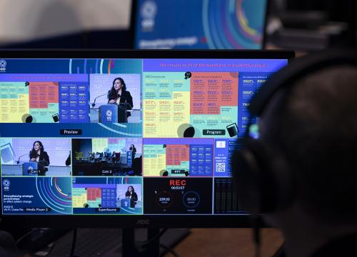 Photograph of monitor during National Report launch, showing Siobhán Nic Fhlannchadha on stage and the results she is reporting on