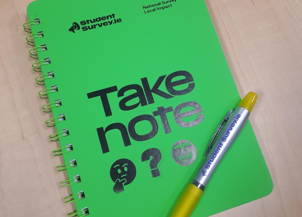 Photograph of a green notebook and silver pen, both with StudentSurvey.ie logos on them